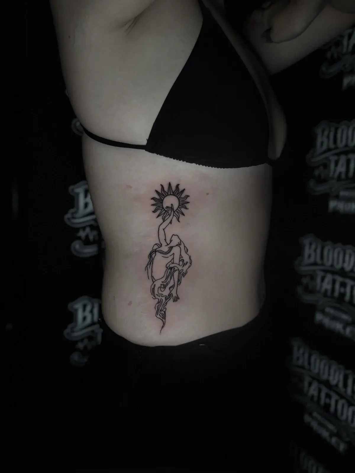 Fine-line bamboo tattoo of a stylized woman reaching toward a radiant sun on the side ribs at Bloodline Tattoo Phuket.