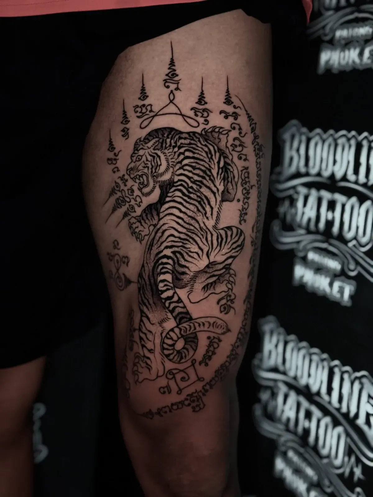 Suea (Tiger) Sak Yant of a snarling tiger with Khom script border and multiple unalomes on the outer thigh at Bloodline Tattoo Phuket.