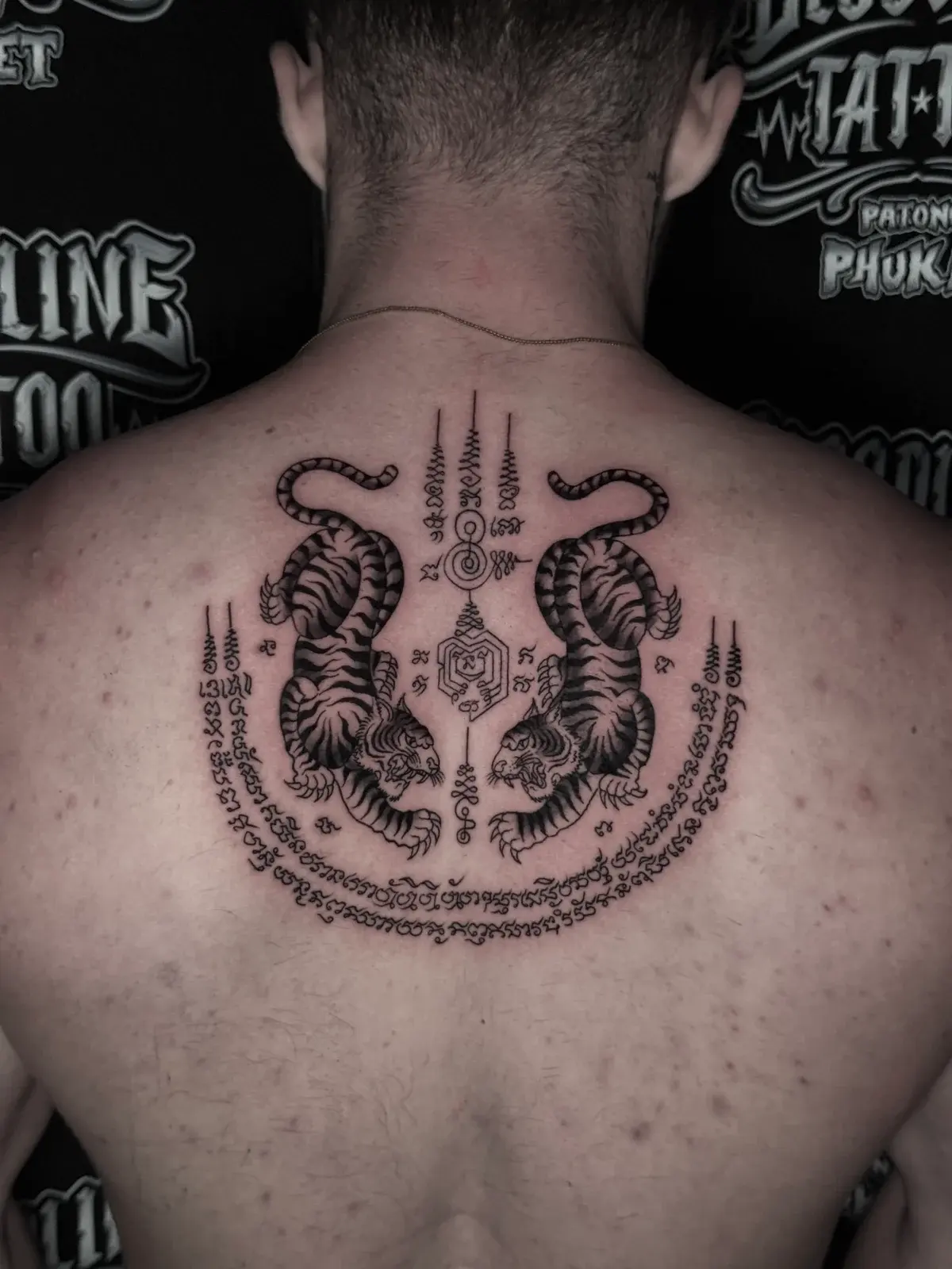 Suea Koo (Twin Tigers) Sak Yant in black and grey with symmetrical tigers, central unalomes, and Khom script crescent on the upper back at Bloodline Tattoo Phuket.