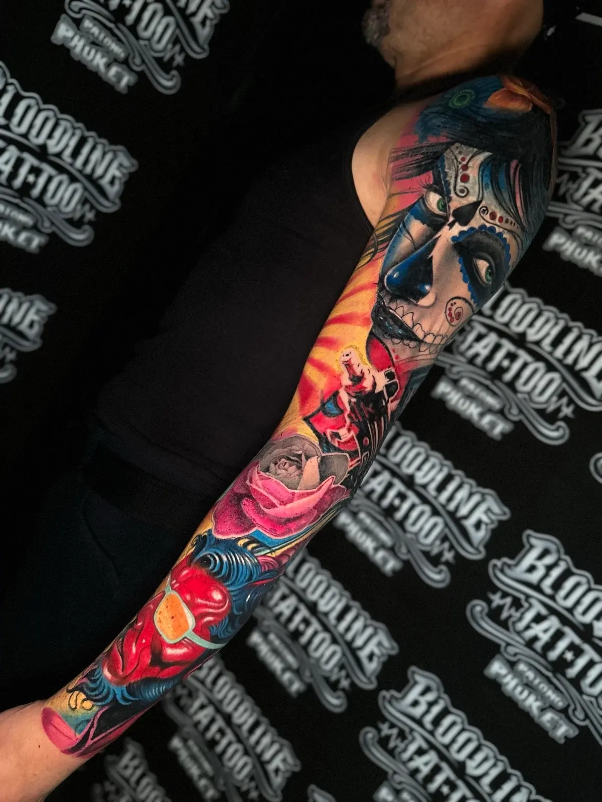 Colour sleeve with a Santa Muerte portrait, a pink rose, and a red demonic face on yellow and blue ornamental backgrounds at Bloodline Tattoo Phuket.