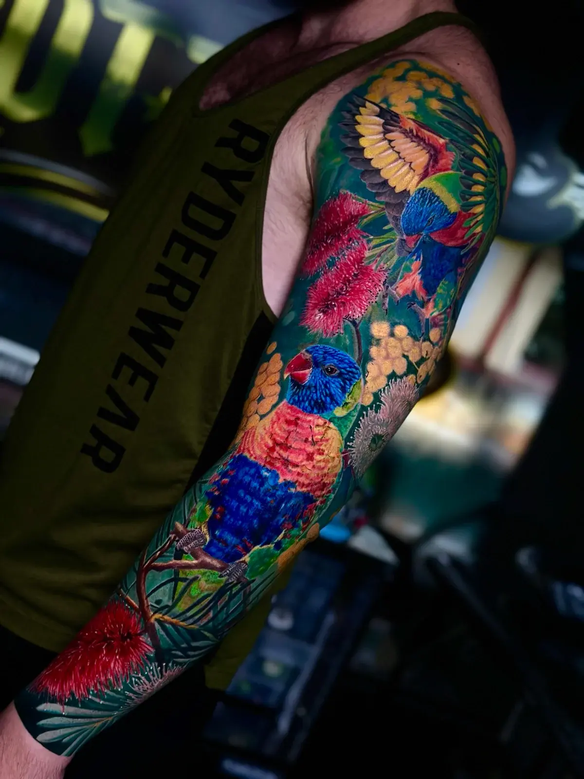 Australian colour sleeve with rainbow lorikeets, red bottlebrush flowers, and golden wattle on dark green foliage at Bloodline Tattoo Phuket.