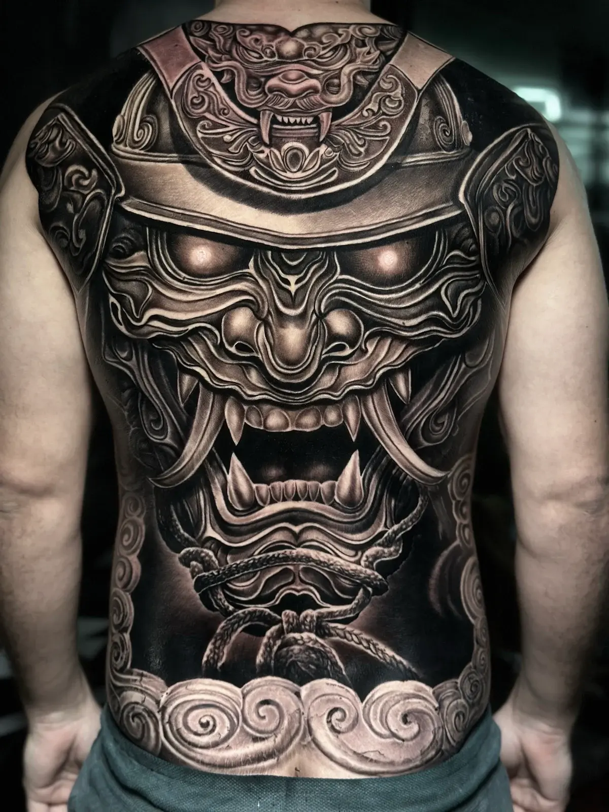 Japanese black and grey full-back samurai menpo with kabuto crest, braided ropes, and stylized clouds on a solid black ground at Bloodline Tattoo Phuket.