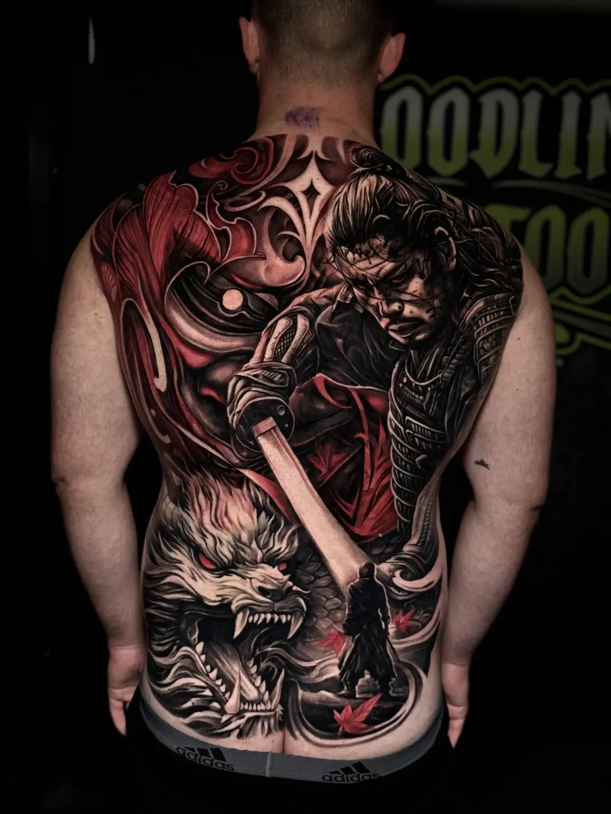 Japanese full-back tattoo with fierce samurai, glowing katana, snarling dragon with red eyes, and red maple leaves at Bloodline Tattoo Phuket.