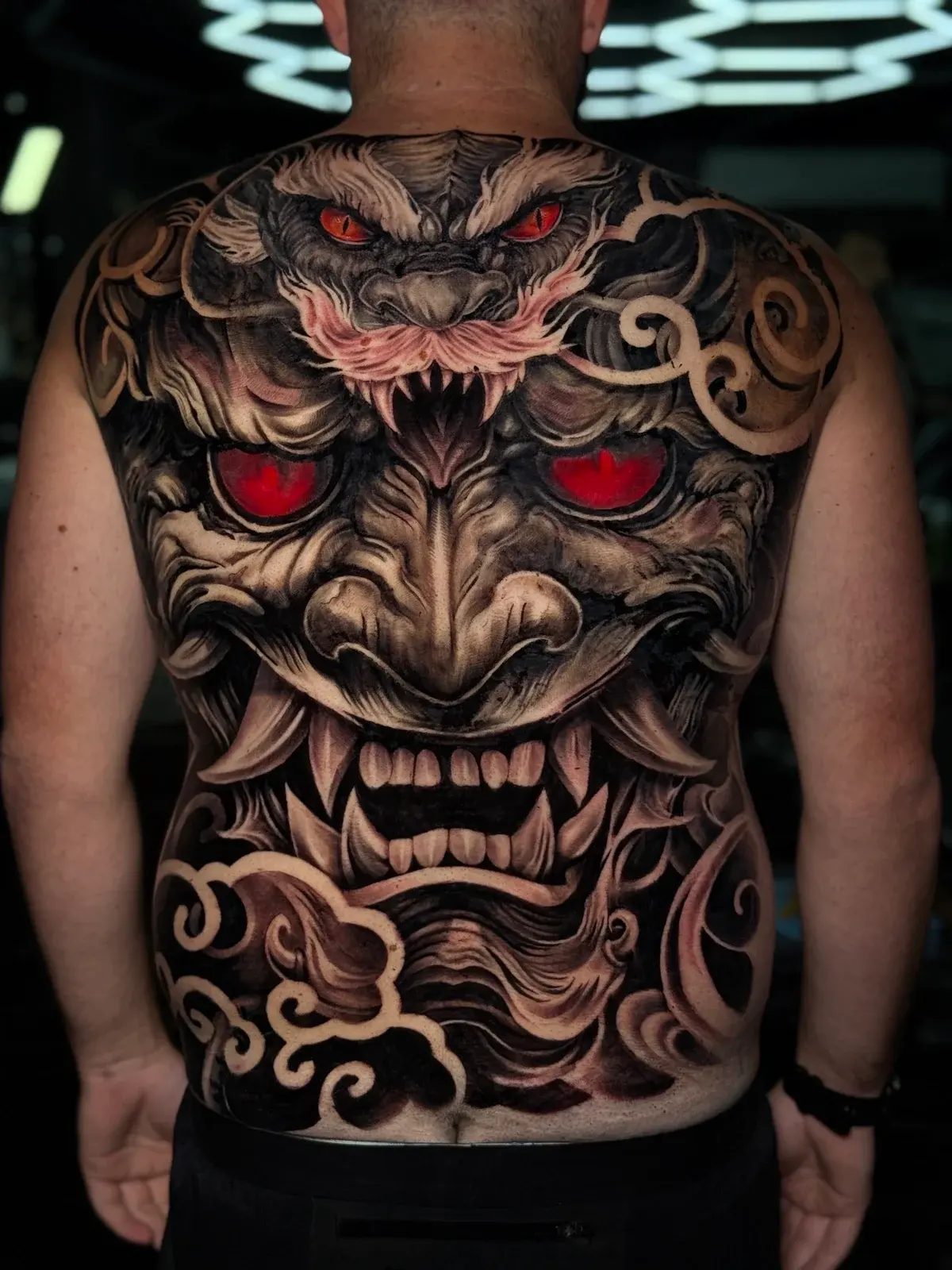 Japanese full-back Hannya mask with glowing red eyes, horned creature crest, and stylized clouds in black and grey at Bloodline Tattoo Phuket.