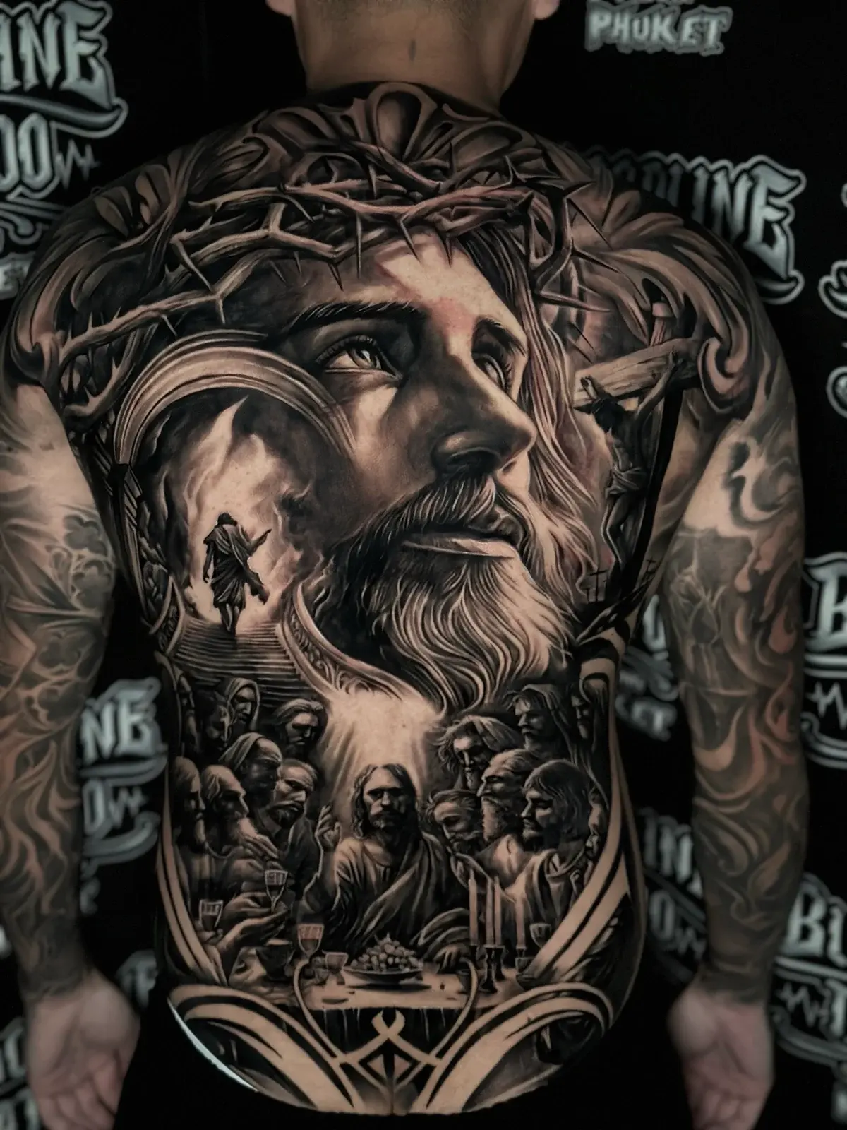Large black and grey realism full back tattoo featuring Jesus with a crown of thorns, the Last Supper, the Crucifixion, and a figure ascending toward light at Bloodline Tattoo Phuket.