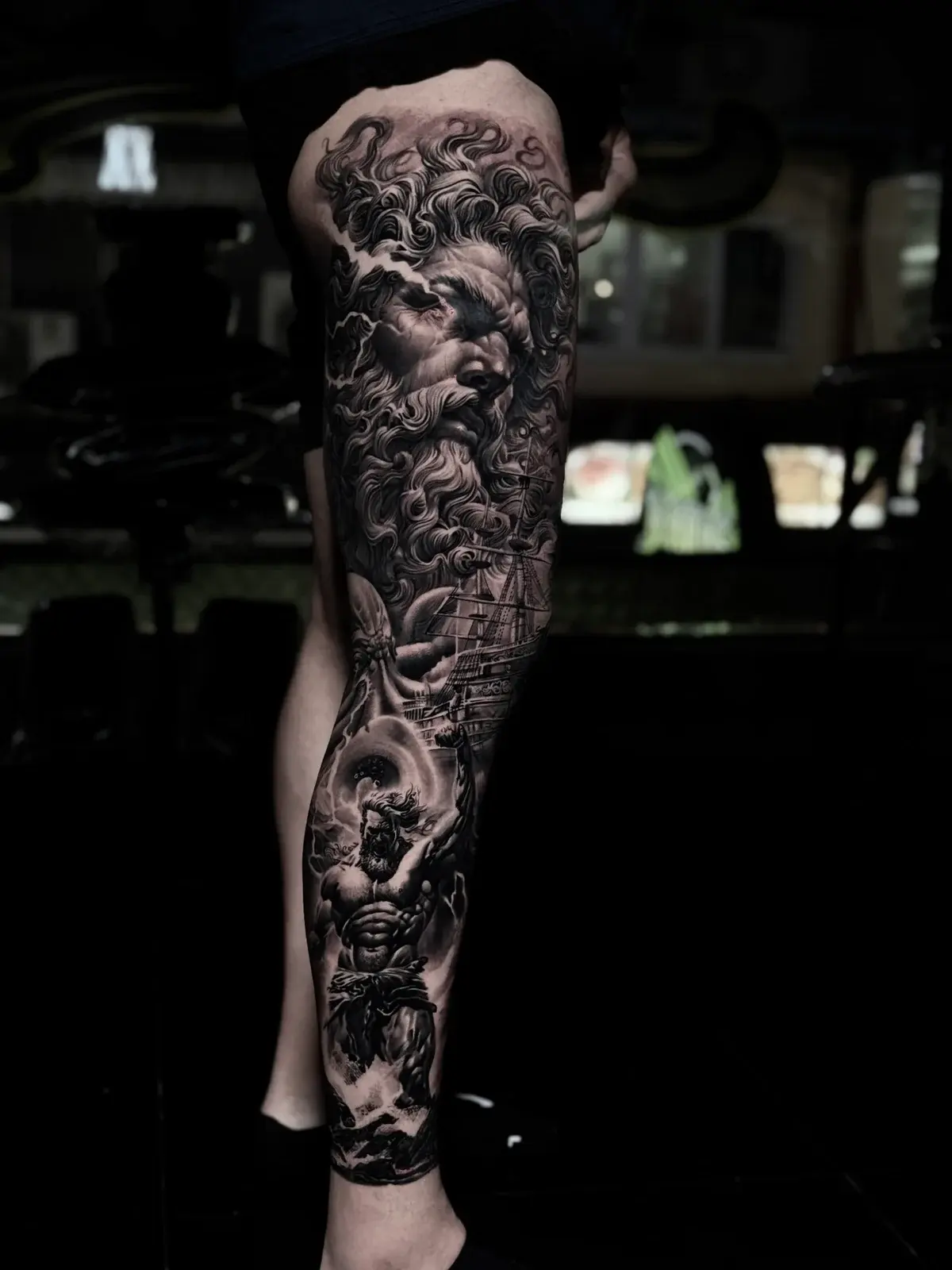 Detailed black and grey realism full leg sleeve tattoo featuring a Greek god portrait with lightning, a sailing galleon, and a muscular figure in stormy waves at Bloodline Tattoo Phuket.