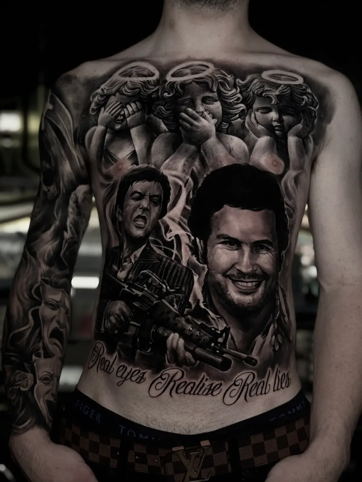 Detailed black and grey realism torso tattoo featuring cherubs, Tony Montana from Scarface, Pablo Escobar, and script reading Real eyes Realise Real lies at Bloodline Tattoo Phuket.