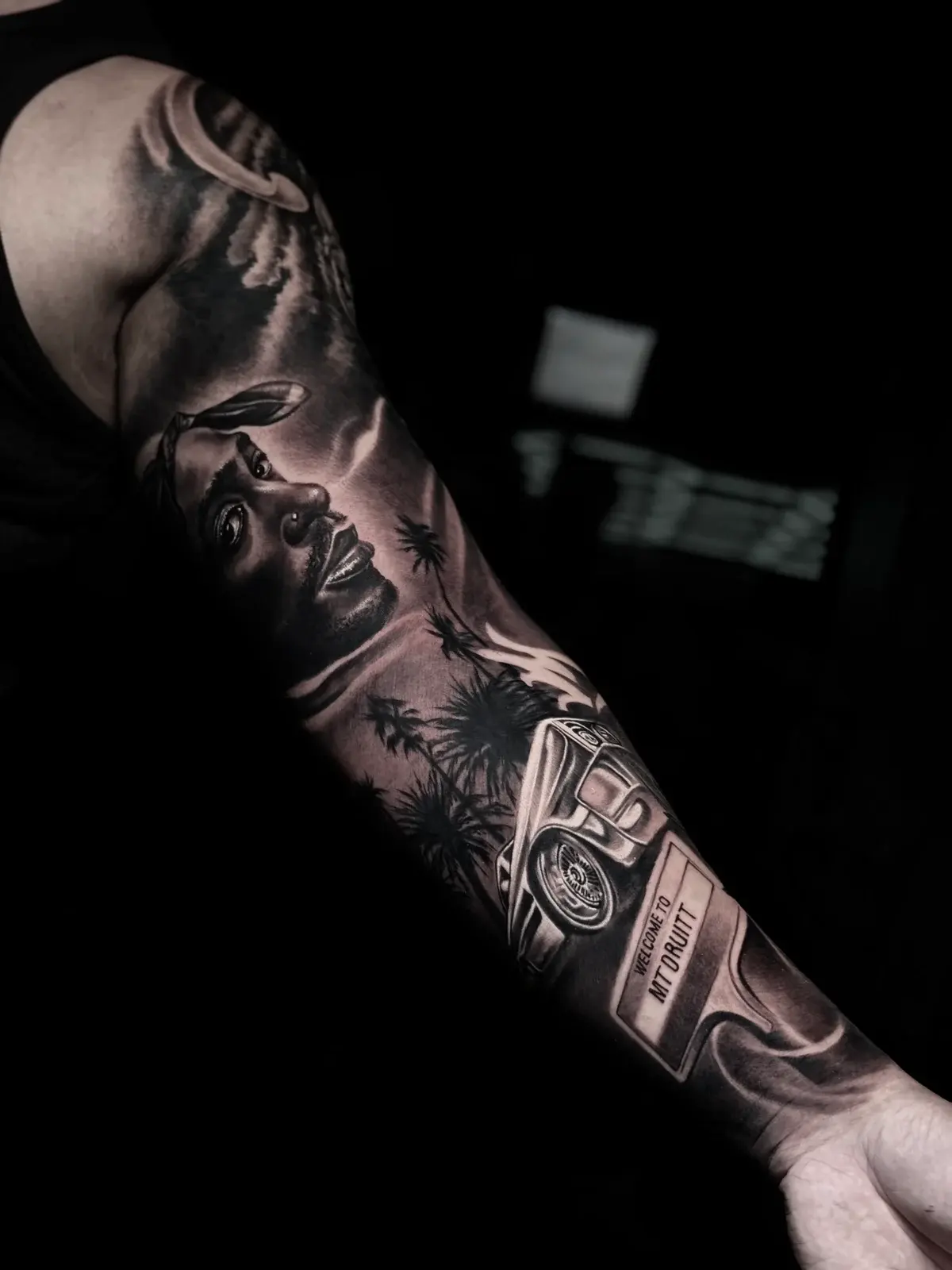 Detailed black and grey realism full sleeve tattoo featuring a Tupac Shakur portrait, palm trees, classic lowrider, and Welcome to Mt Druitt road sign at Bloodline Tattoo Phuket.