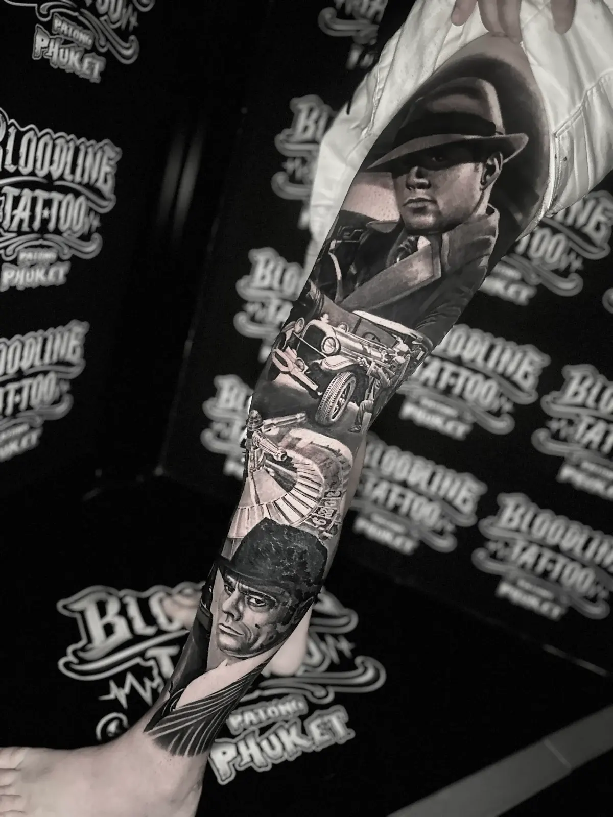 Detailed black and grey realism full leg sleeve tattoo featuring a gangster in a fedora and trench coat, vintage car, roulette wheel, and rugged portrait at Bloodline Tattoo Phuket.